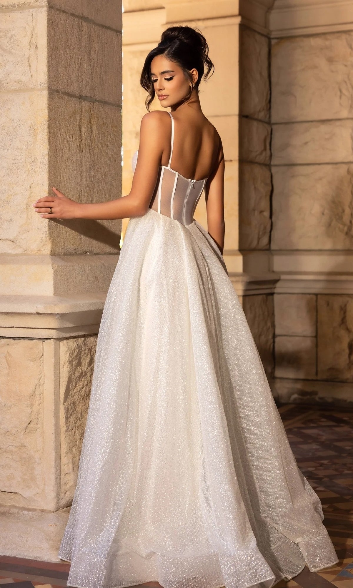 A woman in the Portia and Scarlett PS25312 long prom dress, featuring a glitter A-line silhouette and sheer corset bodice, stands by a stone pillar in an elegant formal setting, looking over her shoulder.