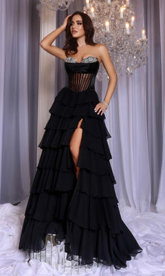 A woman poses indoors under a crystal chandelier wearing the Portia and Scarlett PS25961 long black prom dress, featuring a strapless corset bodice, tiered ruffle skirt with a front slit.