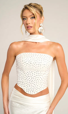 A woman wears the Portia and Scarlett PS26042 long formal dress featuring a white, beaded corset top with silver details, a matching satin skirt, large circular earrings, and a draped white scarf.