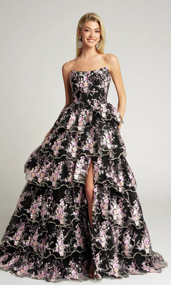 A woman wearing the Portia and Scarlett PS26051 Strapless Floral Ball Gown in black, with embroidered pink and white floral applique, tiered layers, and a front slit, stands and smiles.