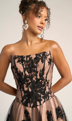 A woman models the Portia and Scarlett PS26061—a long formal a-line dress with a corset bodice and nude and black floral embroidered sheer overlay—against a plain light background.