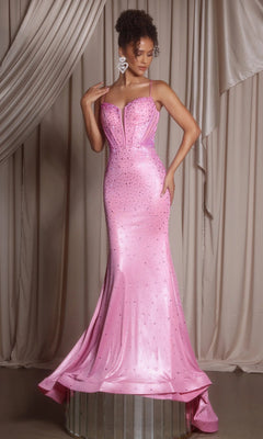 A woman stands on a platform in the pink Portia and Scarlett PS26071 long formal dress, a fitted, floor-length fit-and-flare gown with thin straps and rhinestone embellishments, set against draped beige curtains.