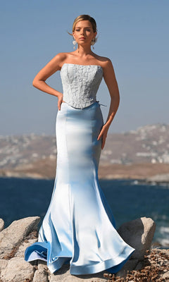 A woman stands on rocks by the water, wearing the Portia and Scarlett PS26092 long formal dress in blue. The strapless gown features a fitted bodice and flared skirt, with hazy hills and a blue sky creating a dreamy backdrop.