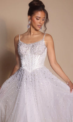 A woman models the Portia and Scarlett PS26208 Corseted Ball Gown in ivory, featuring a beaded boned-corset bodice and thin straps, as she looks down and holds out the skirt against a plain background.