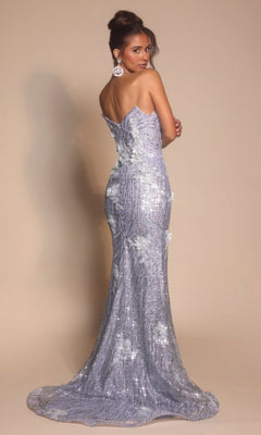 A woman faces backward in the Portia and Scarlett PS26221, a floor-length lavender prom dress with floral details and sparkles, standing against a plain beige background.