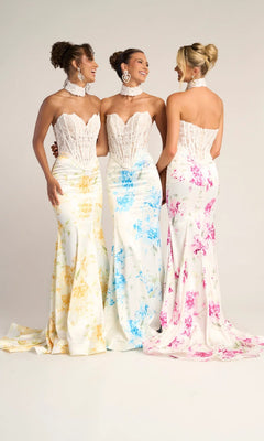 Three women stand together in Portia and Scarlett PS26299 long formal dresses, featuring white strapless lace corset gowns with yellow, blue, and pink floral patterns. They are smiling at each other against a plain background.