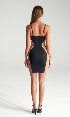 A woman stands facing away in the Short Party Dress PS322B by Portia and Scarlett—a fitted black cut-out dress with nude side panels, thin straps, and black high-heeled sandals—against a plain light background.