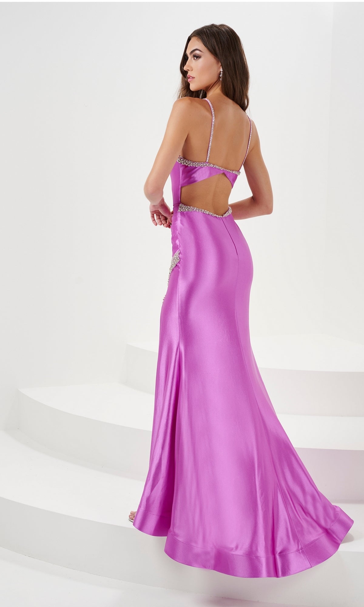 A woman in the Bead-Trimmed Long Mermaid Prom Dress 14173—floor-length, shiny pink with thin straps, beaded trim, and side cutouts—stands on white curved steps, facing away from the camera.