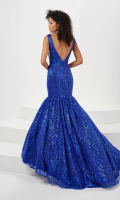 A woman in the Long Prom Dress 14180 by Panoply, a royal blue sleeveless sequin mermaid gown with a deep V-back and open back, stands on white steps facing away, showcasing its floor-length sparkling design.