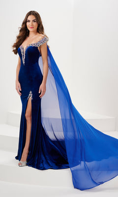 A woman stands on white steps in the Long Prom Dress 14195 by Panoply, an off the shoulder royal blue stretch velvet gown with jeweled details, thigh-high slit, and a long detachable train.