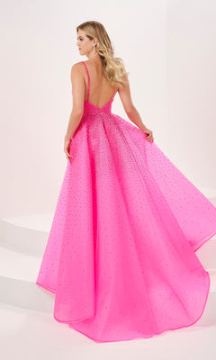 A woman wearing the Long Prom Dress 14201 by Panoply, a bright pink floor-length organza gown with sparkling embellishments and a low v-neckline, stands on white steps holding the sides of her dress.