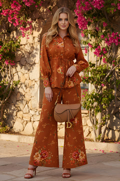 A woman stands outdoors by a stone wall with blooming bougainvillea, wearing the Parker rust floral-printed boho matching set with wide-leg pants and holding a tan handbag.