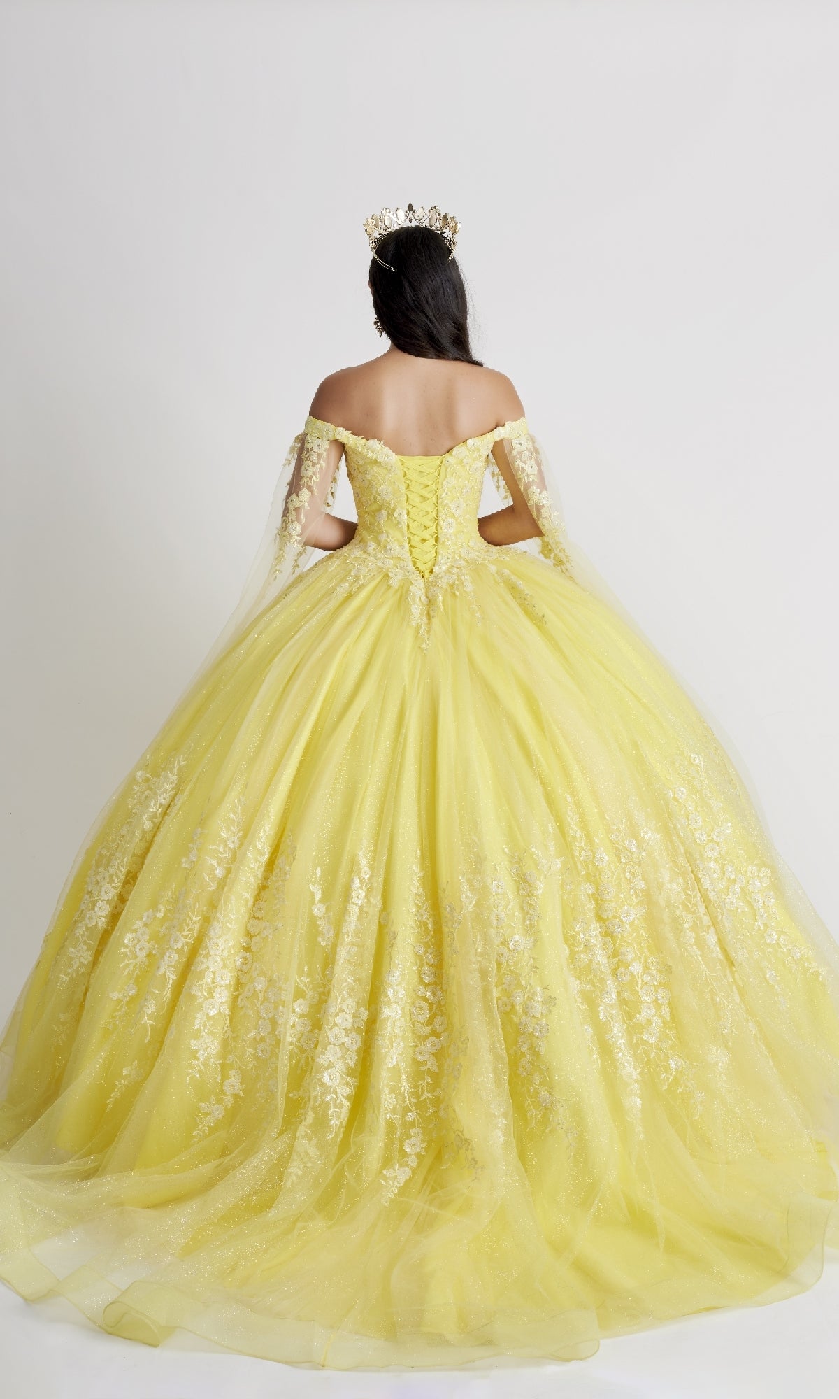 A woman in the Nina Canacci Q109 long quinceañera dress with an off-the-shoulder floral bodice and crown stands facing away against a plain white background.