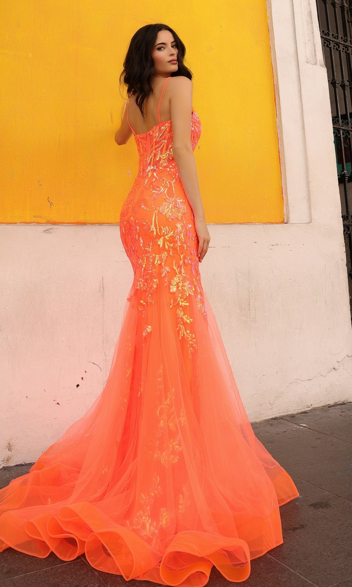 A woman in the Sequin-Lace Long Neon Mermaid Prom Dress Q1390, a bright orange form-fitting gown with floral designs, stands before a yellow and white wall, glancing over her shoulder.