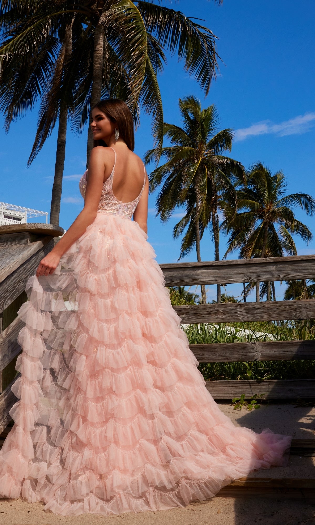 Wearing the Nox Anabel Q1542 Ruffled Long Beaded Prom Dress with a removable overskirt, a woman stands on a wooden walkway among palm trees beneath a clear blue sky.