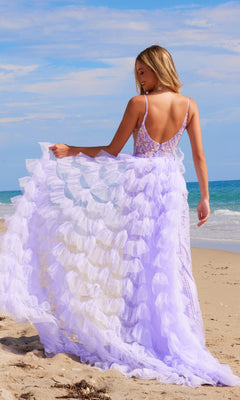 A woman stands on a sandy beach under a blue sky, facing the ocean with her back to the camera, wearing the Nox Anabel Q1542 Ruffled Long Beaded Prom Dress with a removable ruffled overskirt in lavender.