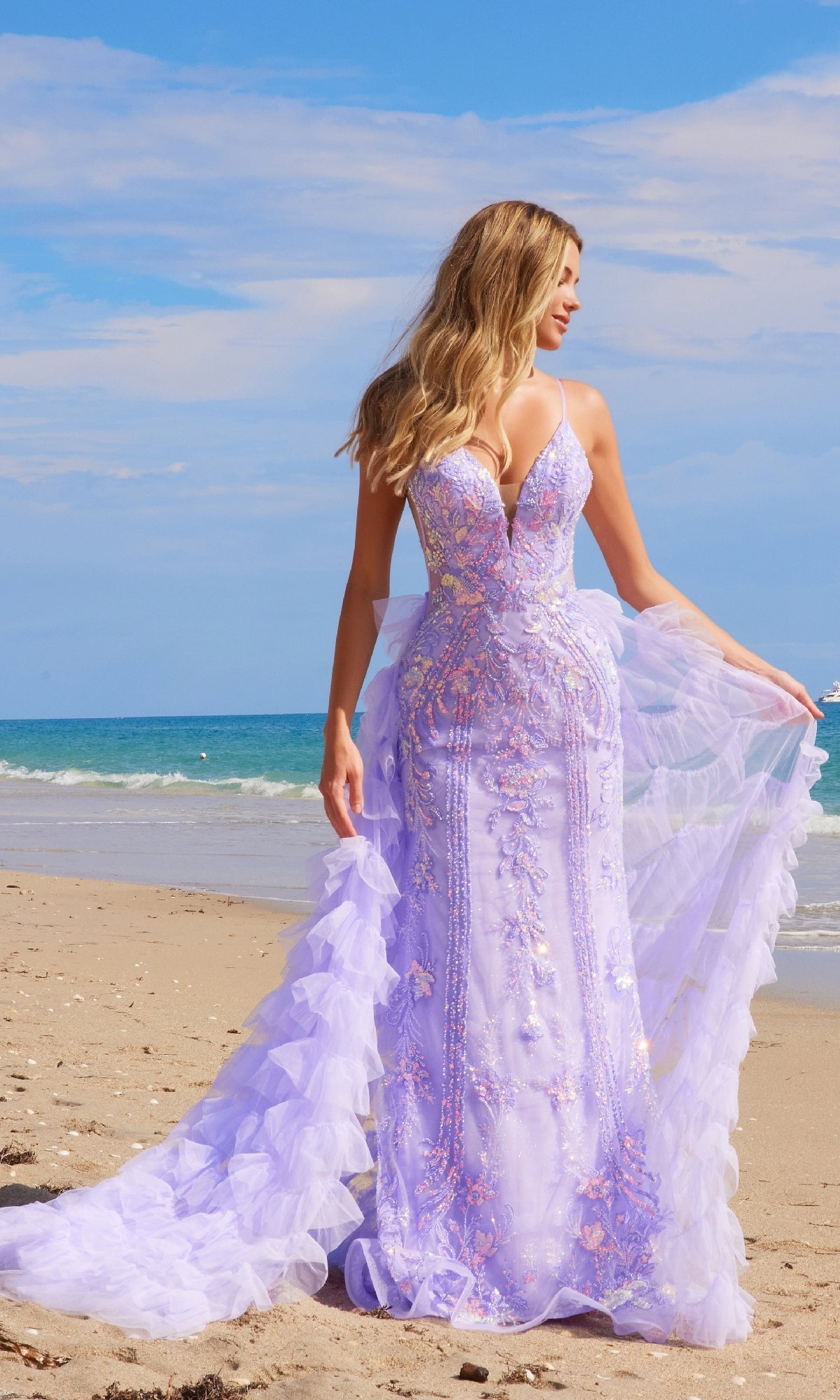A woman in the Nox Anabel Q1542 ruffled long beaded prom dress stands on a sandy beach, with the ocean and blue sky in the background.