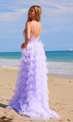 Wearing the Nox Anabel Q1542, a ruffled long beaded prom dress with a removable overskirt, a woman stands on a sandy beach facing the ocean beneath a partly cloudy sky.