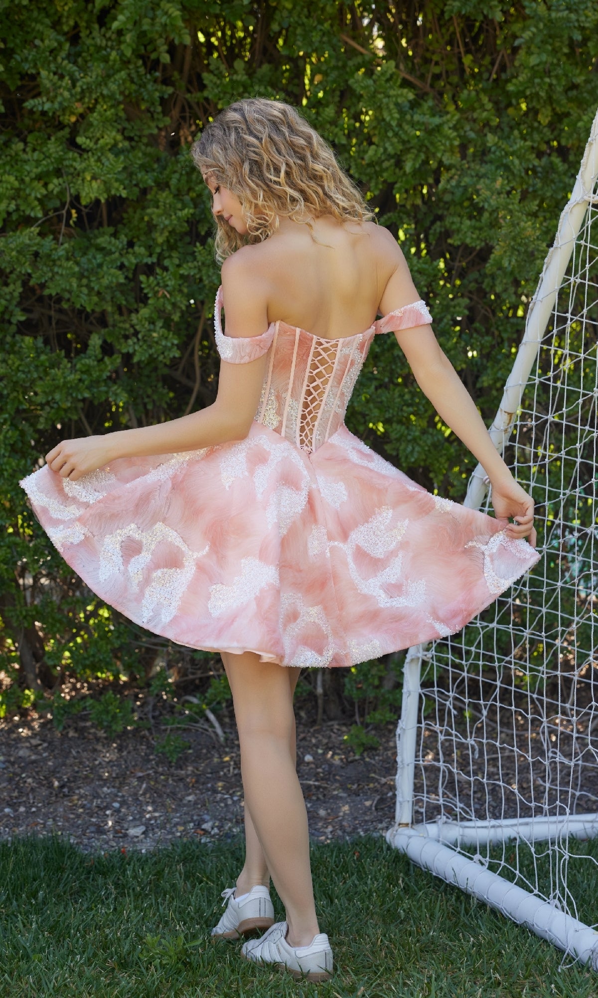 A woman in the Watercolor-Print Homecoming Dress: Nox Anabel Q176 stands on grass near a soccer goal, holding out her skirt and facing away from the camera.