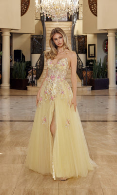 A woman stands indoors wearing the Nox Anabel Q1773 long yellow prom dress with a strapless corset bodice, 3D floral appliques, and a front slit. A staircase and chandelier can be seen in the background.