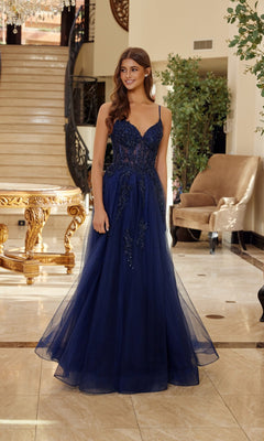 A woman stands indoors in front of a staircase and beige armchair, wearing the Nox Anabel Q1778 long prom dress in navy blue with a fitted bodice and floor-length tulle skirt.