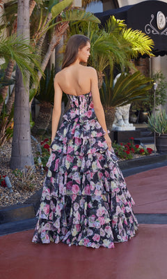 Outdoors, a woman faces away from the camera in the Nox Anabel Q1779 Long Prom Dress, a strapless corset gown with pink, purple, and black florals and chiffon tiered ruffle details.