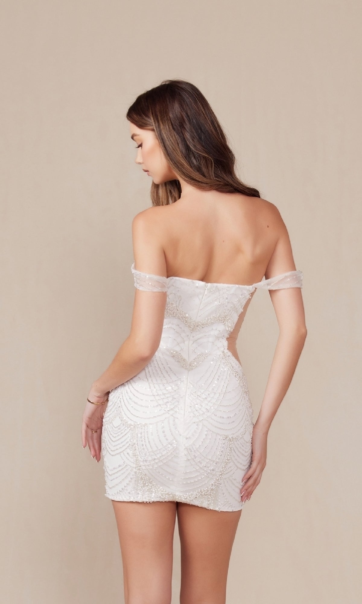 A woman faces away in the Short White Sequin Hoco Dress: Nox Anabel Q839W—an off-the-shoulder, fitted party dress—posing against a plain beige background.