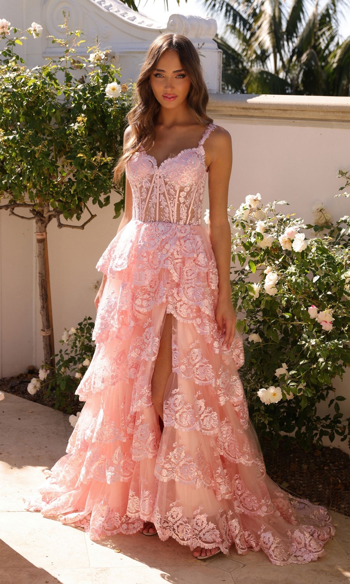 A woman stands outdoors among white roses and greenery, wearing the Nox Anabel Off-the-Shoulder Lace Prom Dress R1299 featuring a tiered pink skirt, front slit, and lace details.