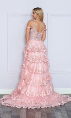 A woman faces away, wearing the Nox Anabel Off-the-Shoulder Lace Prom Dress R1299 in light pink with tiered ruffles and lace details, standing before a white wall and hardwood floor.
