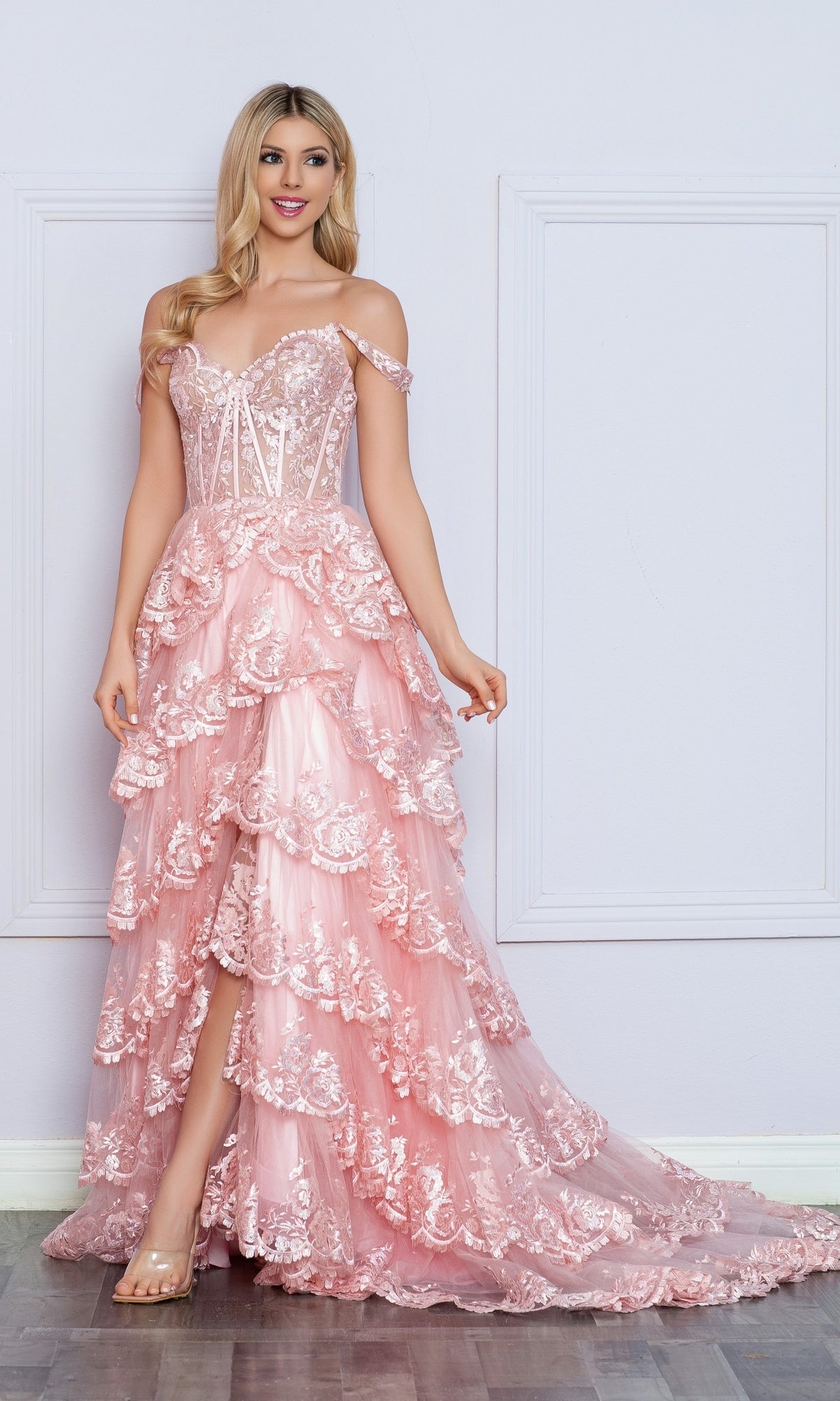 A woman stands against a pale wall, wearing the Nox Anabel Off-the-Shoulder Lace Prom Dress R1299, featuring a tiered pink high-low hemline and a sheer corset bodice.