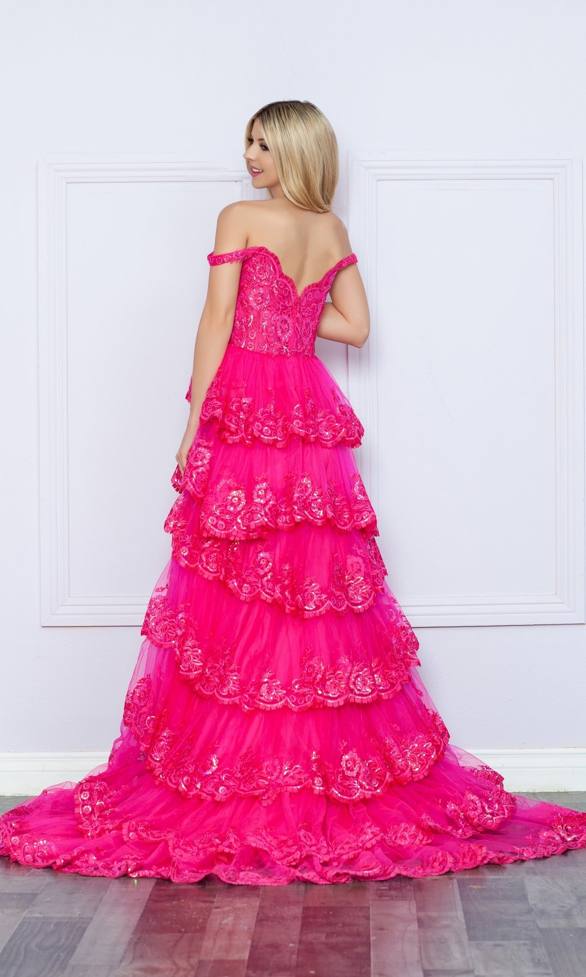 A woman faces away from the camera in the Nox Anabel Off-the-Shoulder Lace Prom Dress R1299, a bright pink tiered gown featuring delicate lace details.