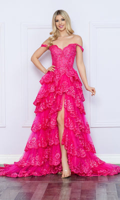 A woman poses indoors in the Nox Anabel Off-the-Shoulder Lace Prom Dress R1299, featuring bright pink lace, tiered layers, floral embroidery, and a high slit against a white wall.