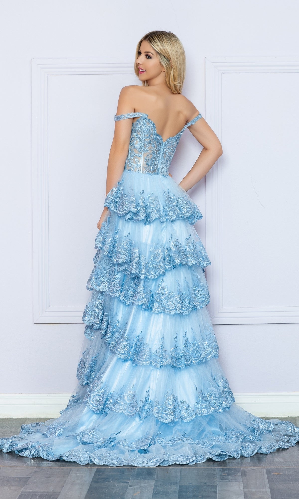 A woman stands with her back to the camera, wearing the Nox Anabel Off-the-Shoulder Lace Prom Dress R1299 in light blue, featuring tiered layers, against a light-colored wall and wooden floor.