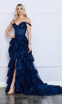 A woman poses in the Nox Anabel Off-the-Shoulder Lace Prom Dress R1299—navy blue, featuring ruffles, a high slit, and a layered skirt—against a light-colored wall and wooden floor.