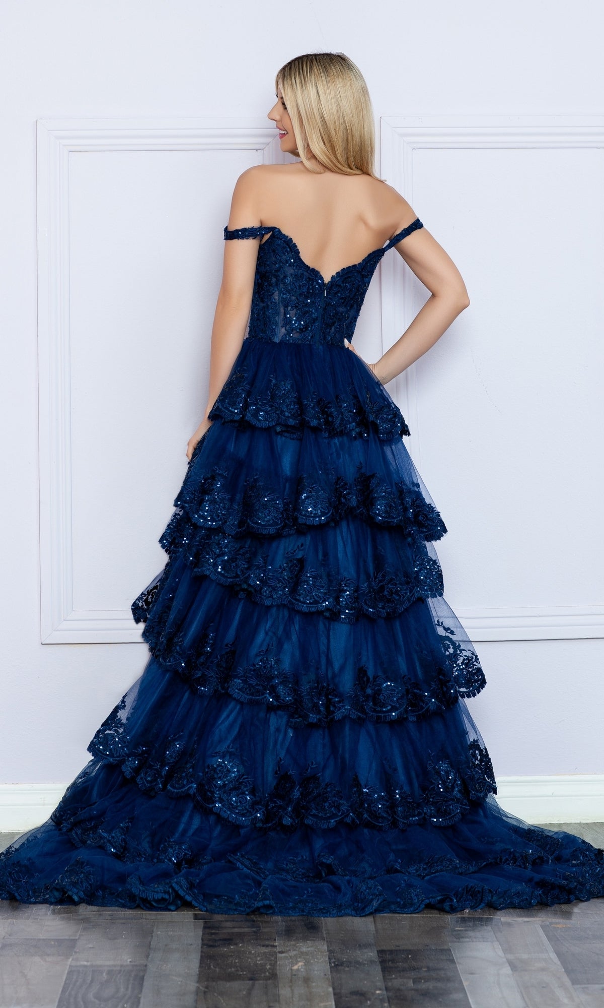 A woman faces away, wearing the Nox Anabel Off-the-Shoulder Lace Prom Dress R1299 in navy blue with tiered layers, sequin accents, and a long train, set against a light wall and wooden floor.