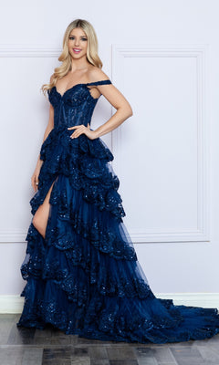 A woman stands indoors against a light-colored wall, wearing the Nox Anabel Off-the-Shoulder Lace Prom Dress R1299 in dark blue, featuring ruffles, sequins, and a thigh-high slit.