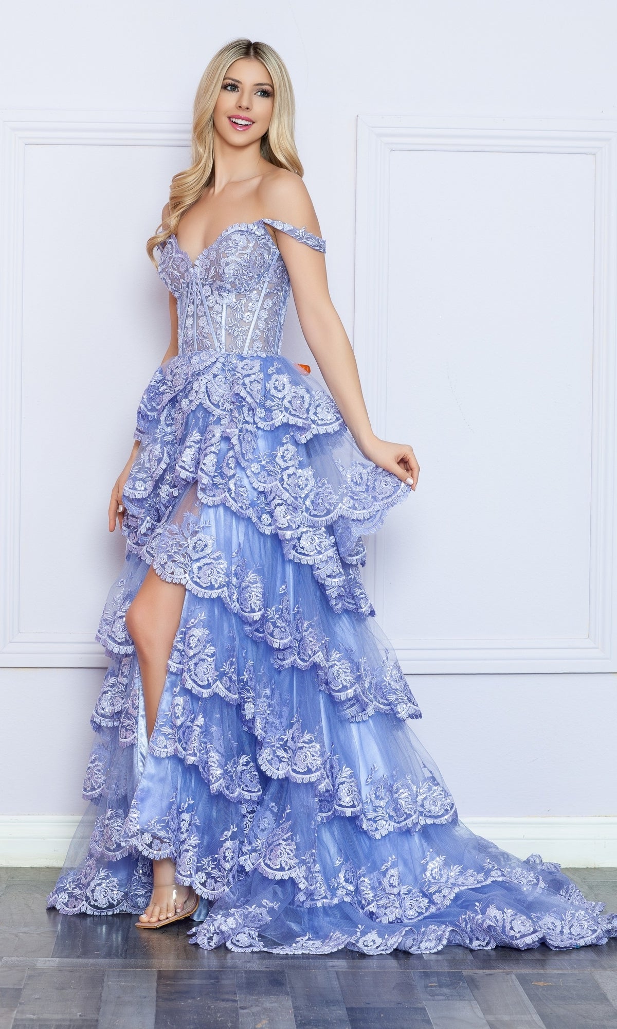 A woman wears the Nox Anabel Off-the-Shoulder Lace Prom Dress R1299 in light purple, featuring ruffled tiers and a sheer corset bodice, while standing against a white paneled wall on a wooden floor.