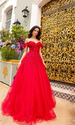 A woman in the Off-the-Shoulder Long Tulle Prom Ball Gown R1303 stands before an ornate gold wrought iron gate and a building decorated with floral accents.