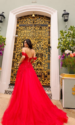 A woman in the Off-the-Shoulder Long Tulle Prom Ball Gown R1303, bright red and flowing, stands before an ornate gold and black door framed by elegant floral arrangements.