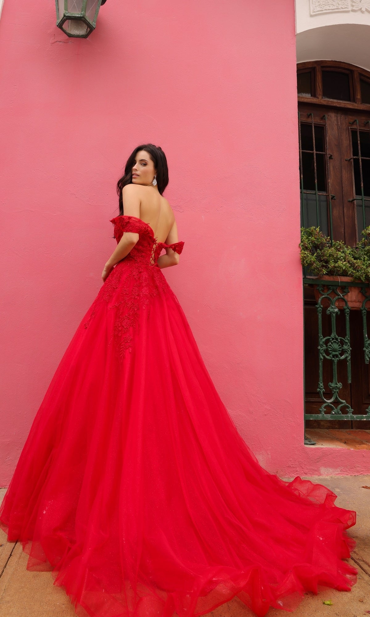 A woman in an Off-the-Shoulder Long Tulle Prom Ball Gown R1303 stands before a pink wall, glancing over her shoulder; her elegant red dress showcases a chic off-the-shoulder style.