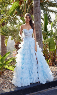 A woman stands outdoors among palm trees and lush greenery in the Nox Anabel R1688, a light blue strapless ruffled prom ball gown with a sheer corset bodice.