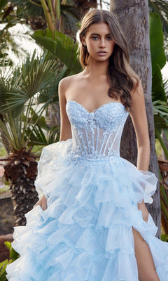A woman stands outdoors near trees and greenery, wearing the Nox Anabel R1688 Strapless Ruffled Prom Ball Gown in light blue, featuring a long skirt and sheer bodice.