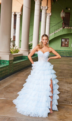 A woman in the Nox Anabel R1688 Strapless Ruffled Prom Ball Gown with a thigh-high slit stands on a tiled walkway, framed by columns and a green building in the background.