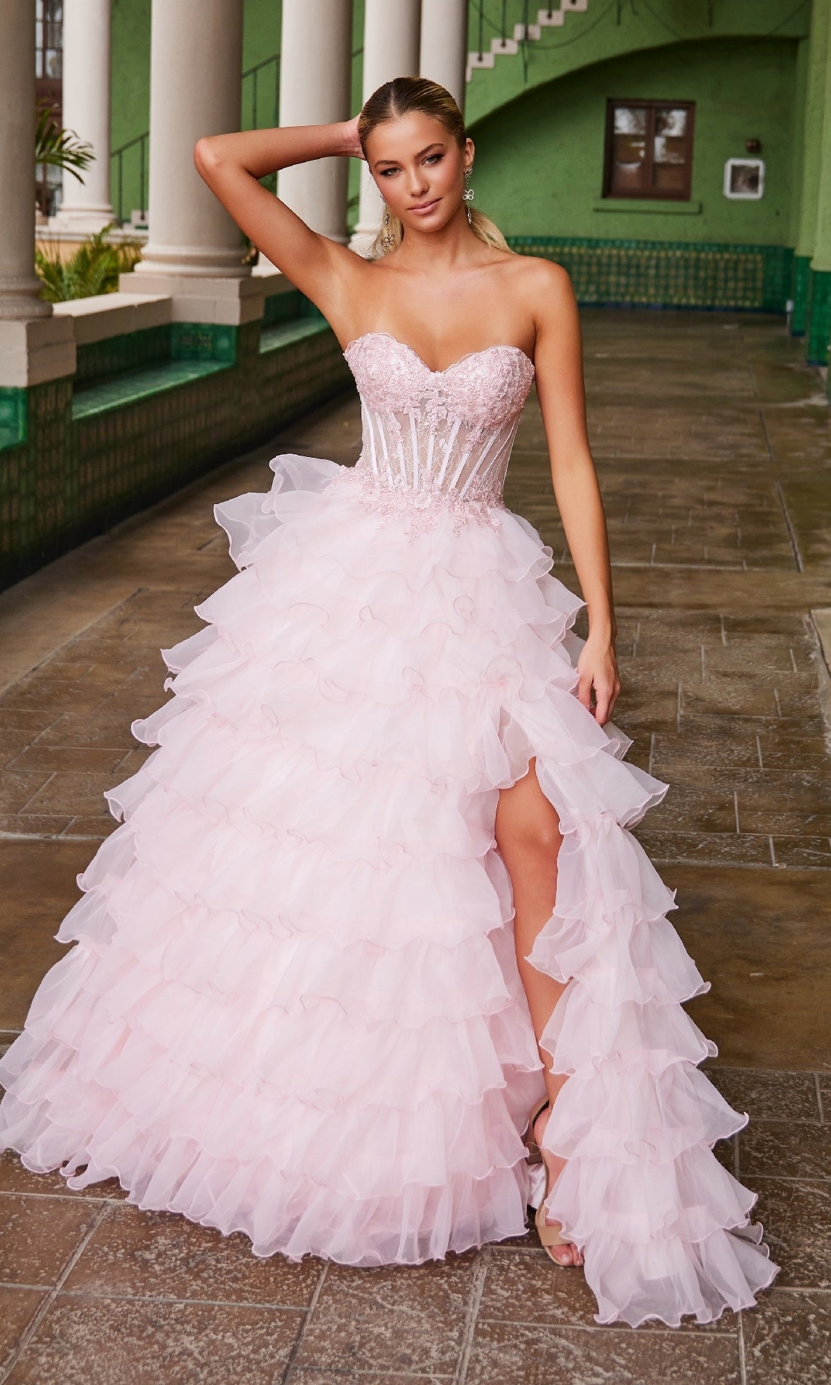 A woman poses in an outdoor corridor wearing the Nox Anabel R1688, a strapless pink ruffled prom ball gown with a fitted beaded bodice and high-slit skirt.