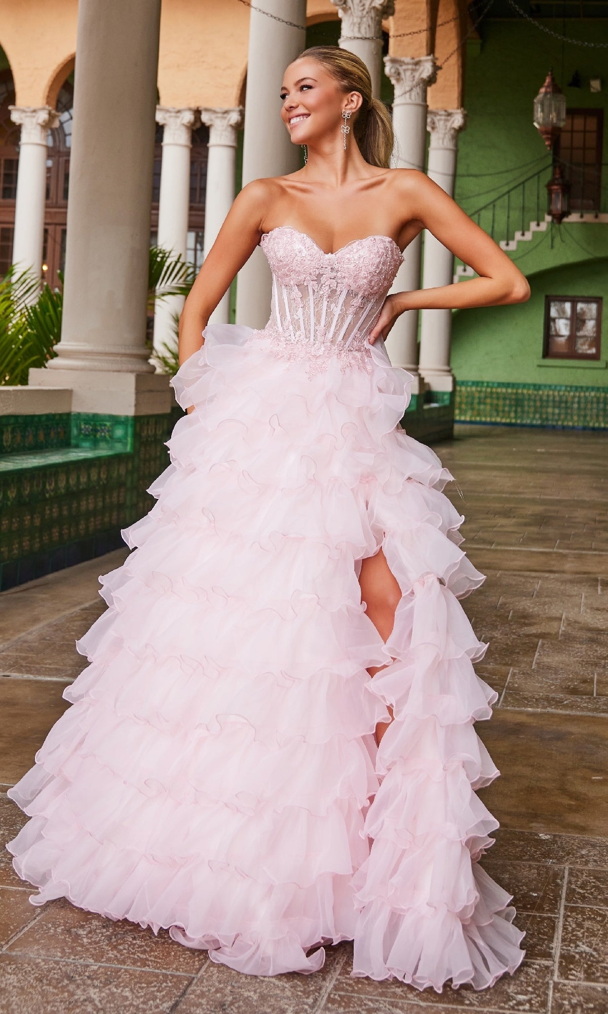 A woman wearing the Nox Anabel R1688, a strapless ruffled prom ball gown in light pink with a fitted beaded bodice, stands in an ornate outdoor hallway.