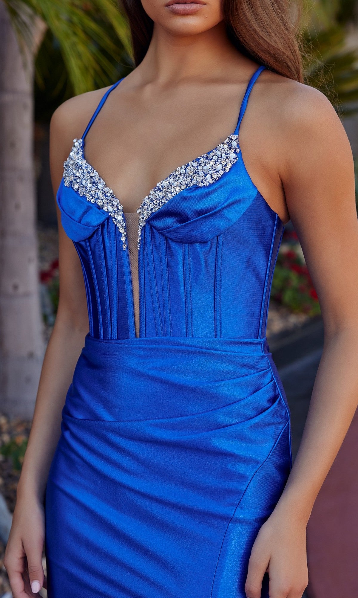 A woman in the Nox Anabel R1689 Lace-Up Long Corset Prom Dress, a fitted royal blue satin gown featuring thin straps, a lace-up open back, and rhinestone accents along the neckline.