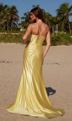 A woman stands on a sandy beach, facing away from the camera, wearing the Nox Anabel R1689 Lace-Up Long Corset Prom Dress in yellow. Palm trees sway gently in the background.