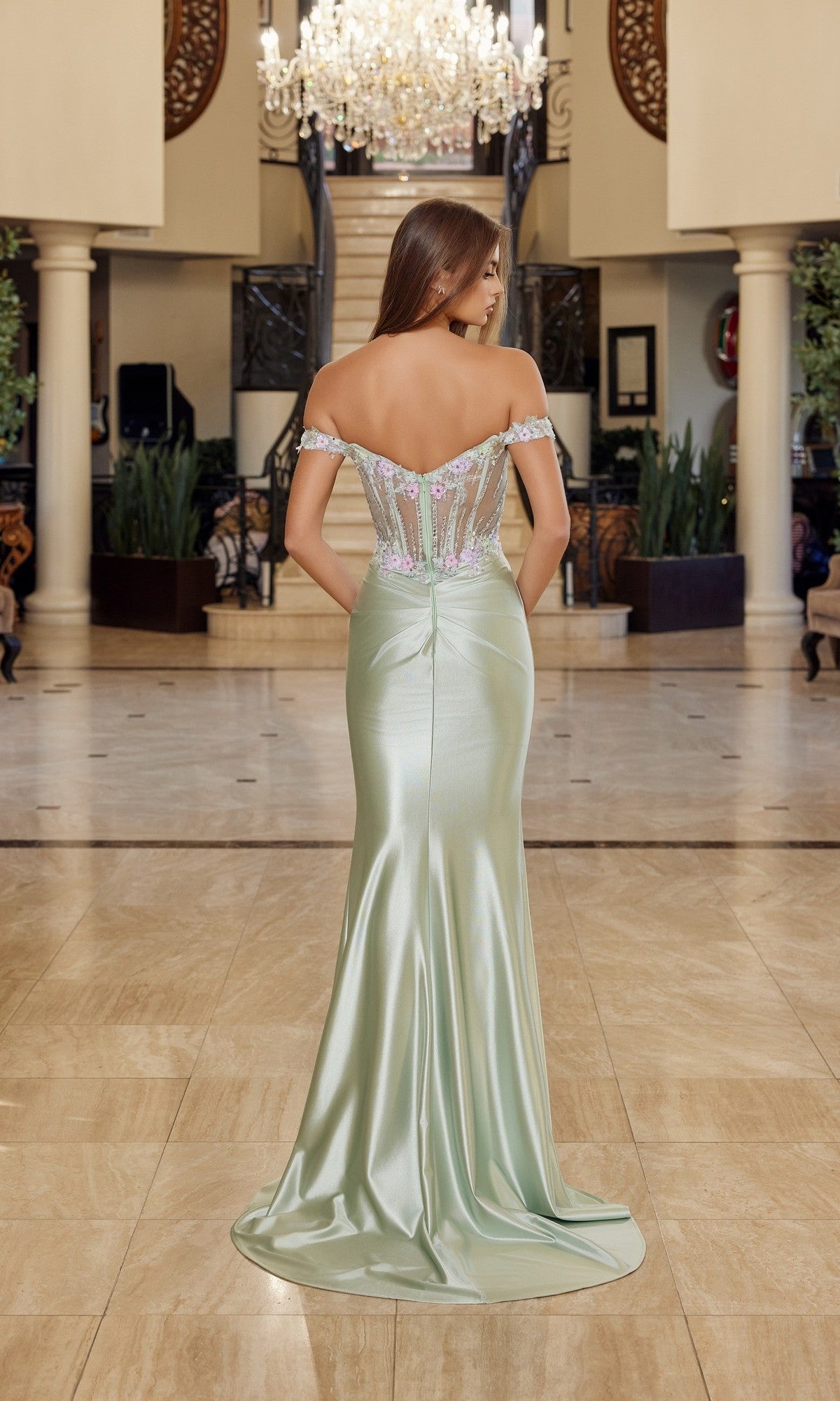 A woman stands indoors in the Nox Anabel R1814 long prom dress, featuring a light green off-the-shoulder design, floor-length satin sheath skirt, and delicate floral details, with her back to the camera.