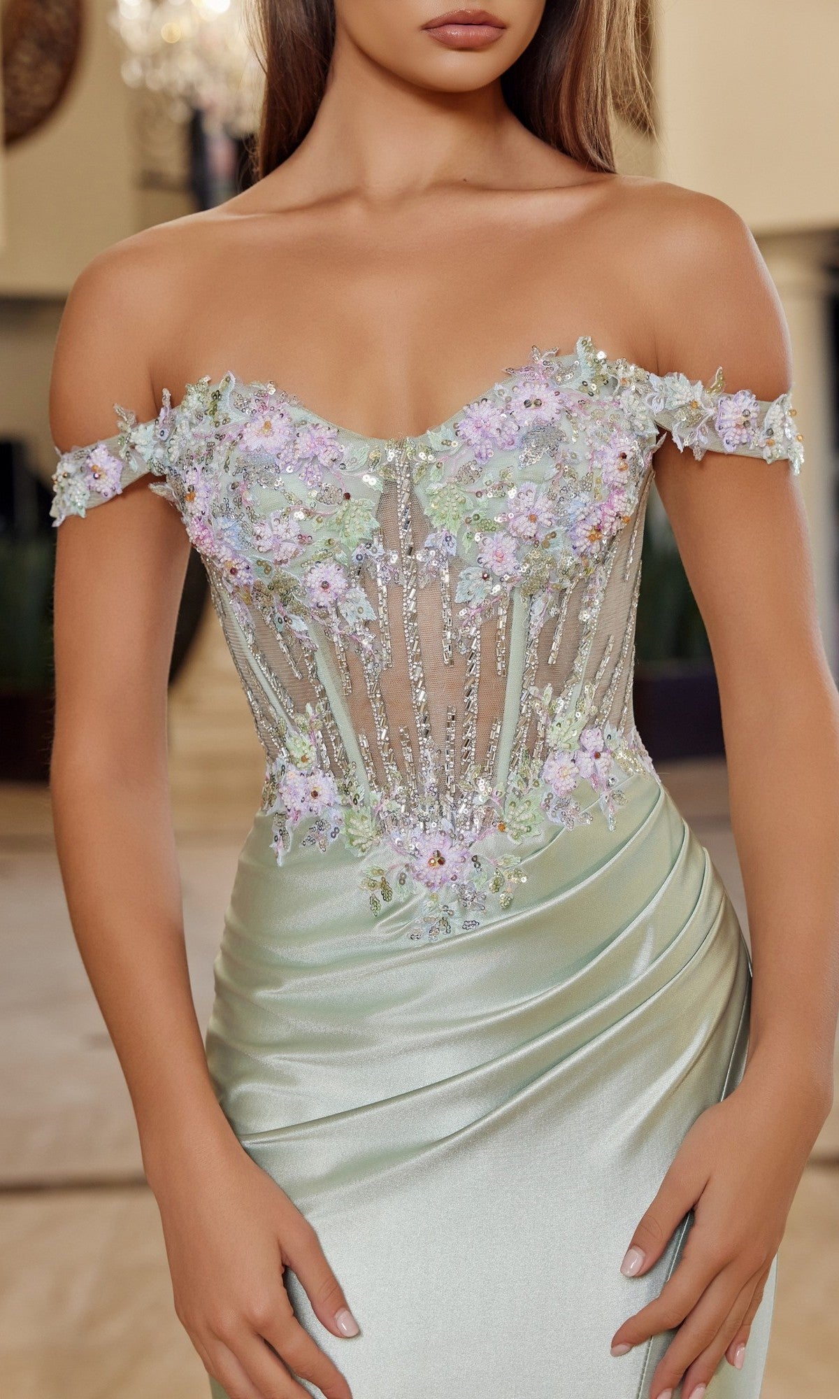 Woman wearing the Nox Anabel R1814 long prom dress, featuring an off-the-shoulder design with a sheer, floral-embroidered bodice and a light green satin sheath skirt, standing indoors.