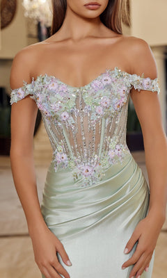 Woman wearing the Nox Anabel R1814 long prom dress, featuring an off-the-shoulder design with a sheer, floral-embroidered bodice and a light green satin sheath skirt, standing indoors.